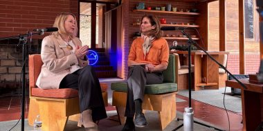 Two women seated on wooden chairs, engaged in a conversation in a room with microphones